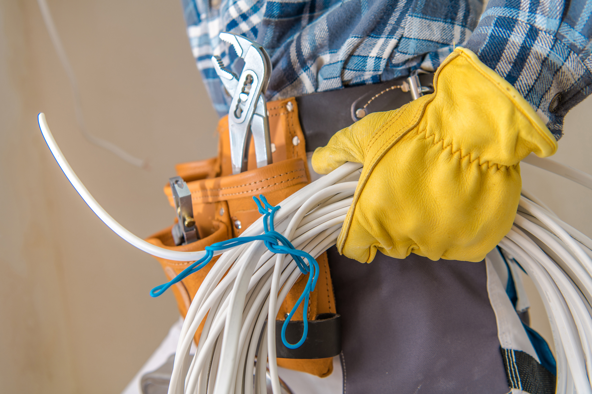 A person wearing yellow gloves and a plaid shirt holds a bundle of white cables. A tool belt with pliers and a screwdriver is visible.