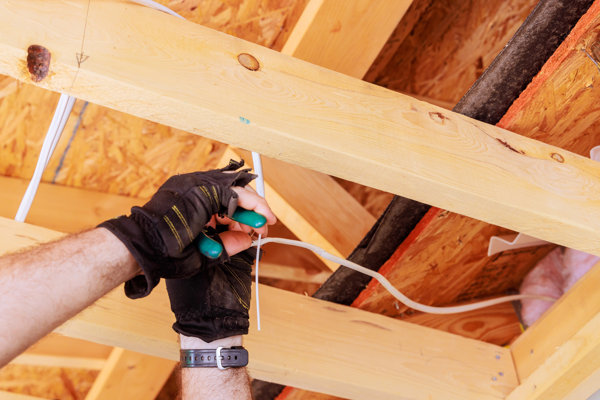 Person wearing black gloves uses pliers to cut a wire within exposed wooden beams of a ceiling.