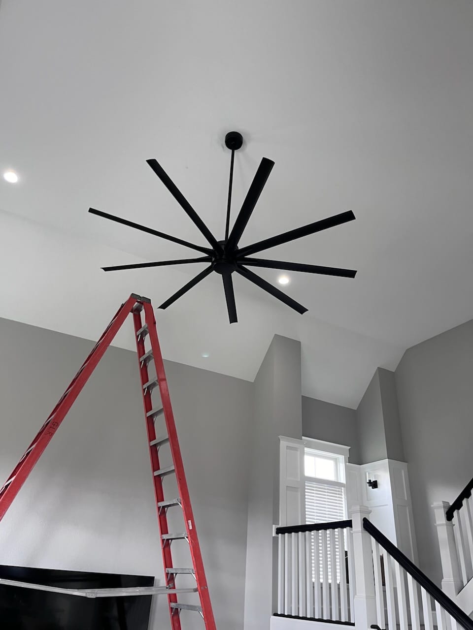 A large black ceiling fan with multiple blades is installed on a white ceiling. A red ladder stands below it in a room with gray walls and white trim.